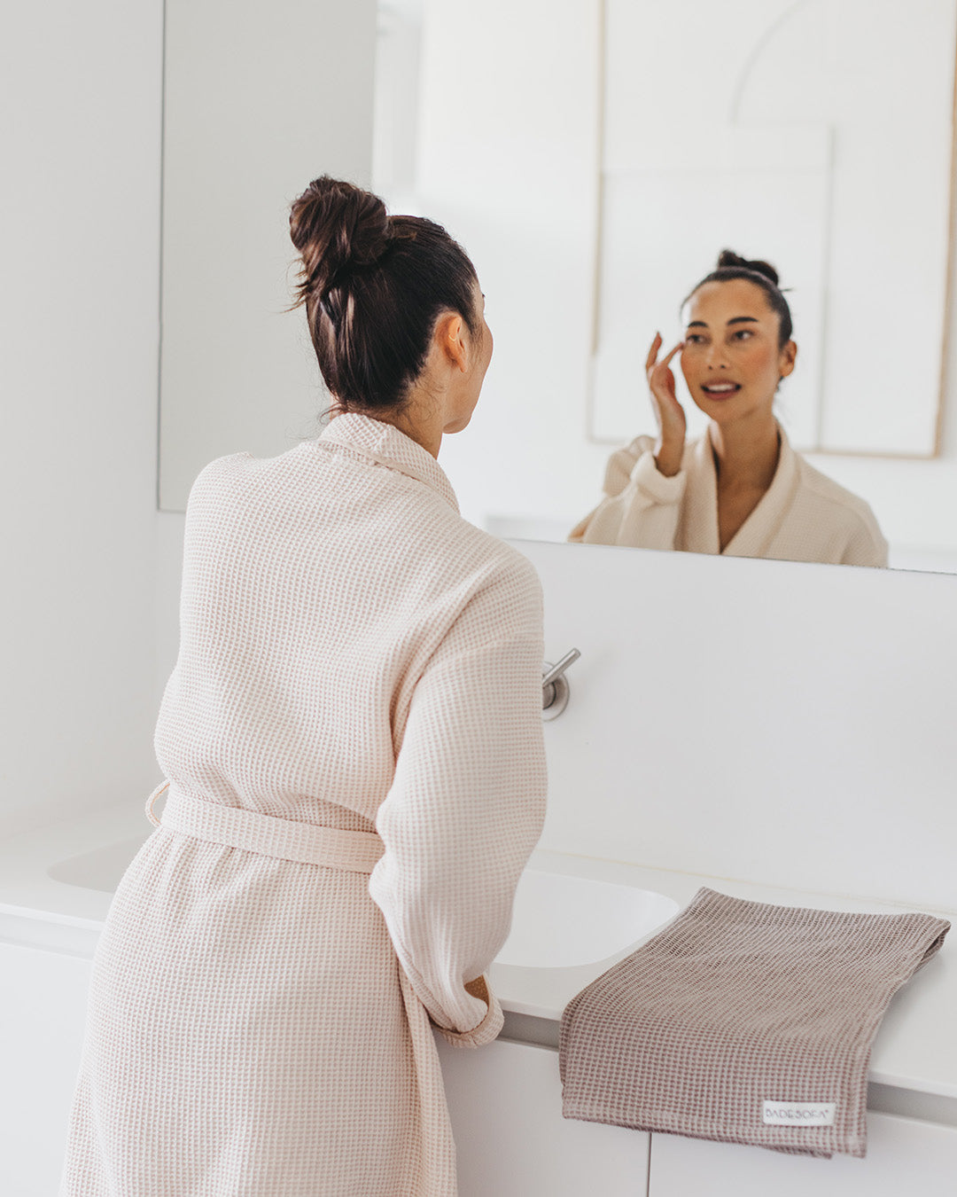 Frau steht im Bademantel (Farbe Perlmutt) vor dem Spiegel, neben ihr liegt ein Saunatuch in Taupe.