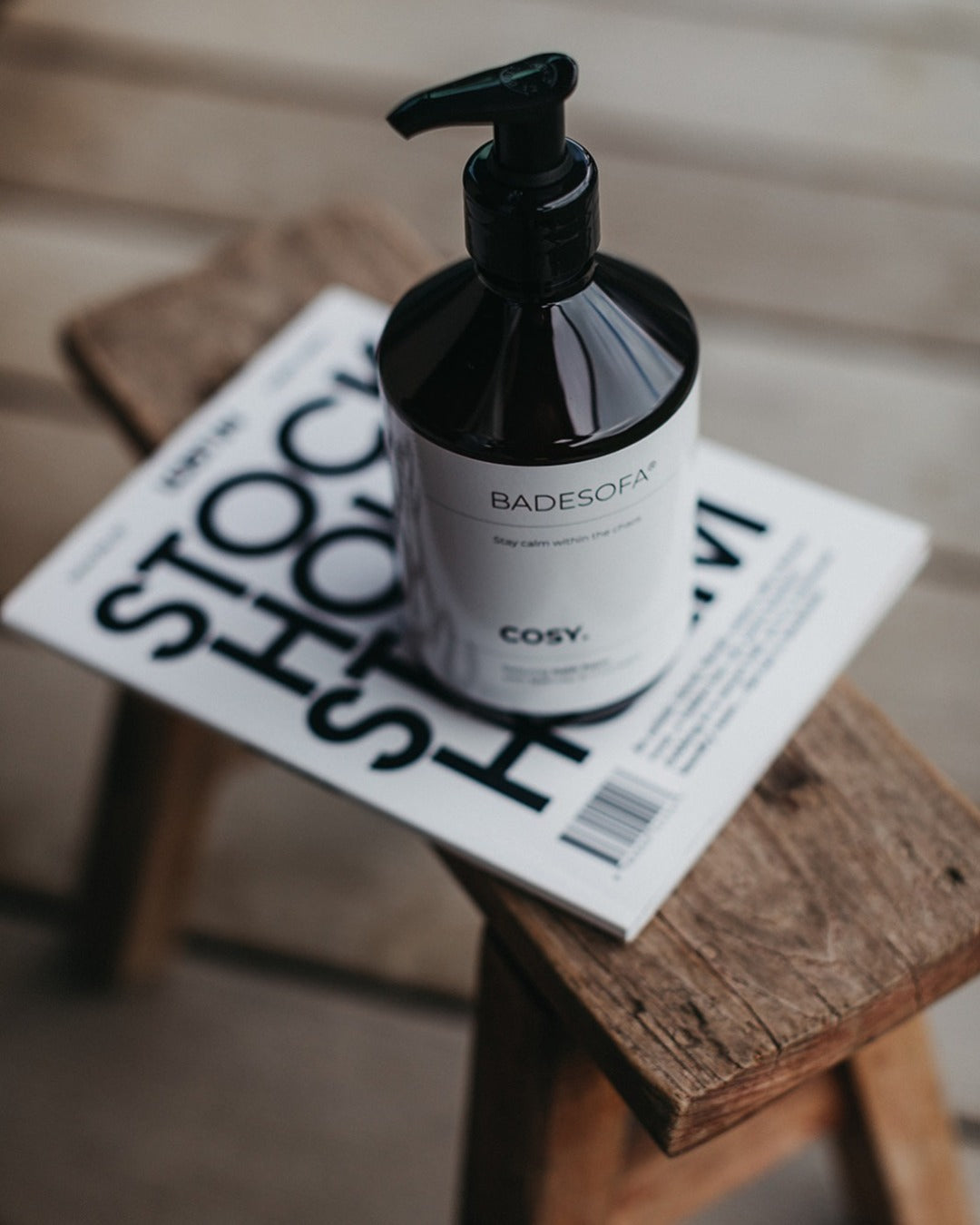 Auf einem Holzhocker steht eine Flasche Badesofa Badeschaum Cosy dekoriert auf einem Magazin.